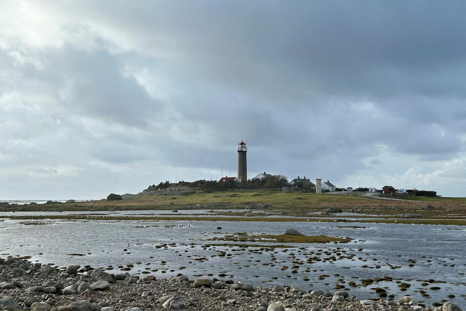 Lista fyr sett fra stranden, med grunt vann i forgrunnen og truende skyer på himmelen.